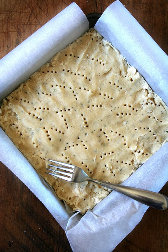 A square baking dish filled with rosemary shortbread dough, pricked with a fork. 