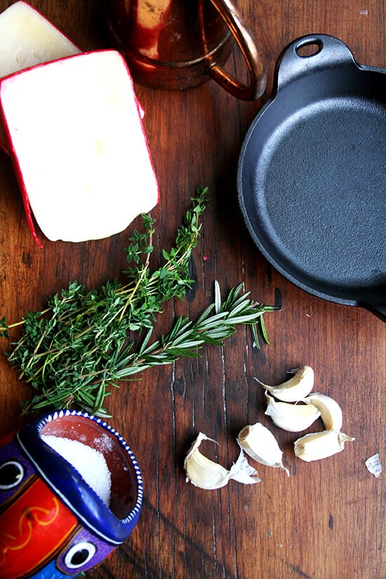 ingredients for baked fontina on a board: fontina, rosemary, thyme, garlic, olive oil, salt.