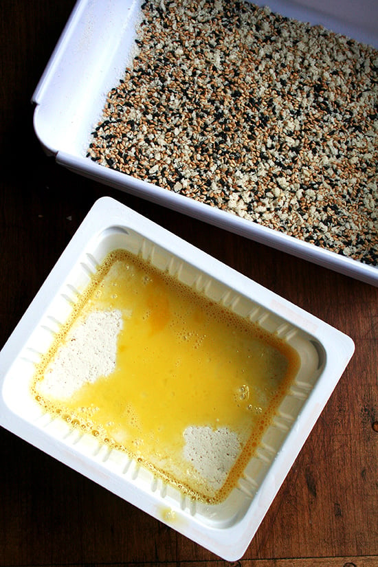 A tray with sesame seeds and panko aside a tray with beaten egg.