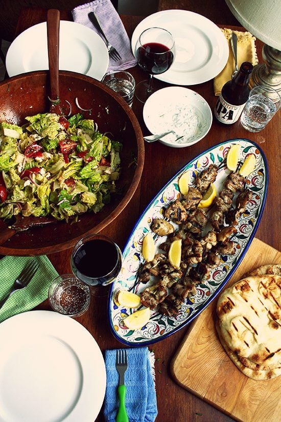 Overhead shot of the dinner table: Greek chicken souvlaki, Greek salad, tzatziki, grilled pita.