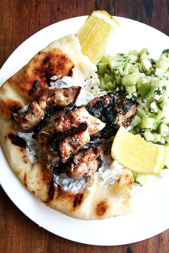 A platter of chicken souvlaki with lemon, tzatziki, cucumber salad, and pita.