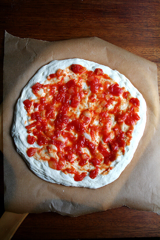 Stretched round of Lahey pizza dough on a pizza peel topped with sauce.