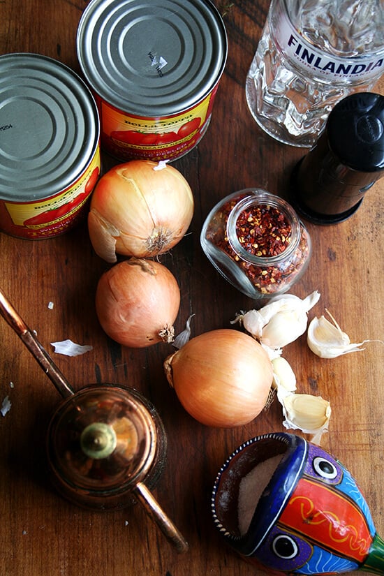 Ingredients gathered on wooden surface: onions, crushed red pepper, pepper grinder, canned tomatoes, vodka.
