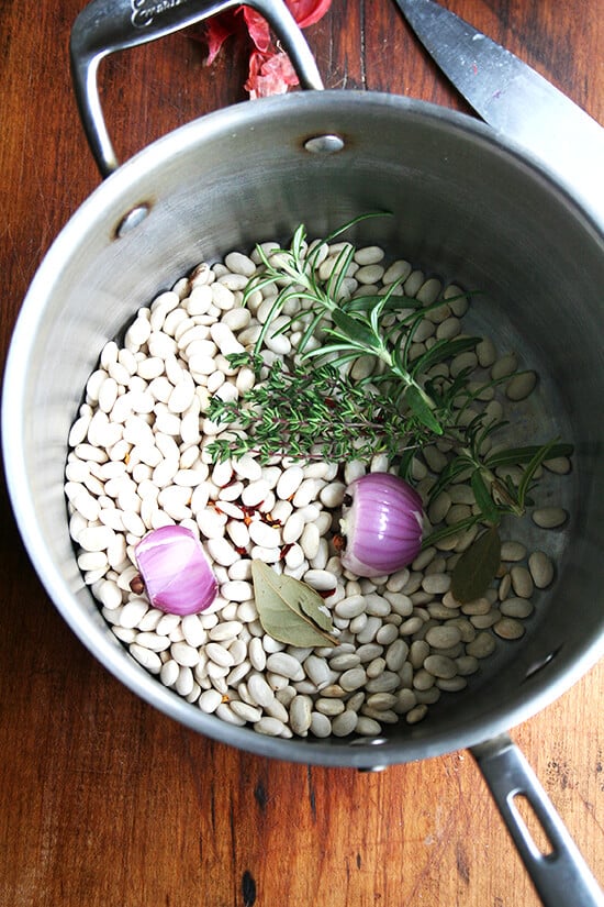 A pot filled with dried white beans, herbs, a bay leaf, and half a shallot. 