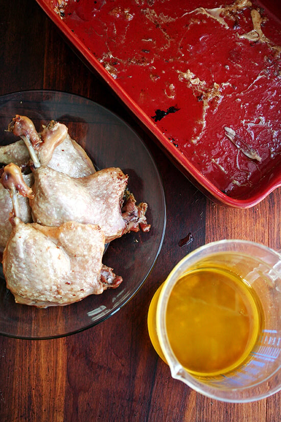 Four duck leg confits on a plate aside a liquid measure filled with rendered fat. 