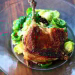 A duck leg confit standing up on a plate supported by a mound of salad greens.