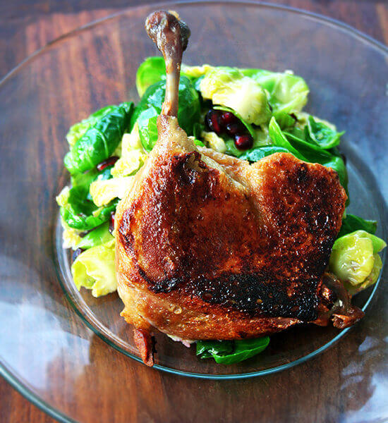 A duck leg confit standing up on a plate supported by a mound of salad greens.