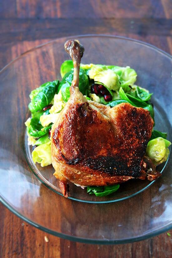 A duck leg confit standing up on a plate supported by a mound of salad greens.