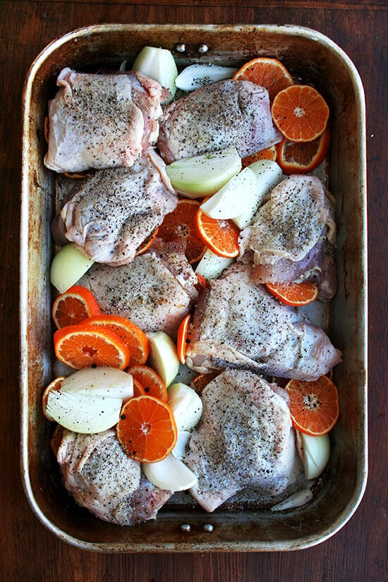 A roasting pan filled with seasoned chicken and raw onions and clementines.