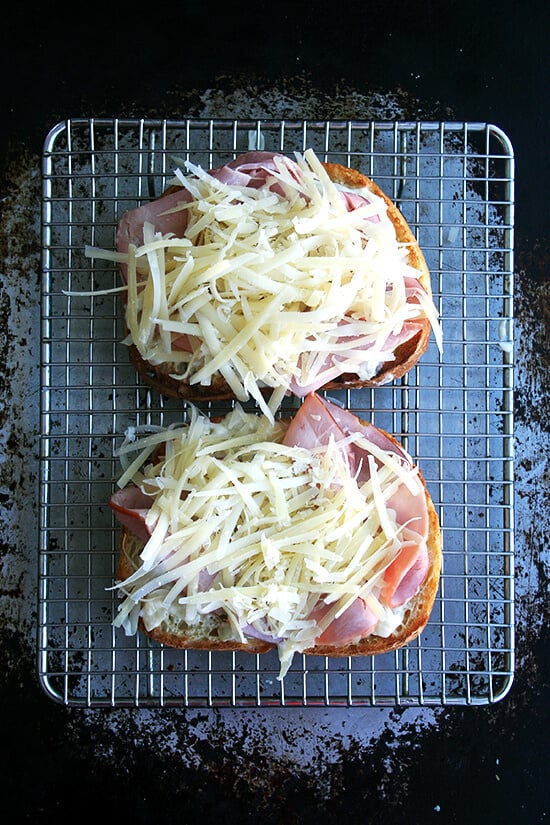 Croque monsieur in the making: Two slices of toast topped with béchamel, ham, and cheese on a rack set on a sheet pan.