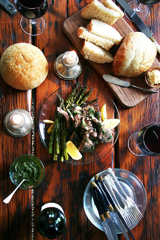 An overhead shot of a dinner with homemade bread, mint sauce, broiled lamb chops, and red wine.