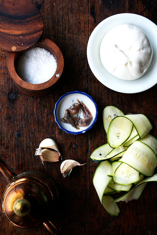A table of ingredients for the zucchini anchovy pizza: anchovies, salt, zucchini, garlic, burrata, and olive oil. 