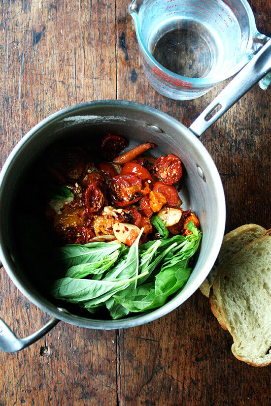 A saucepan filled with roasted vegetables and basil aside a liquid measure with water and a few slices of stale bread.