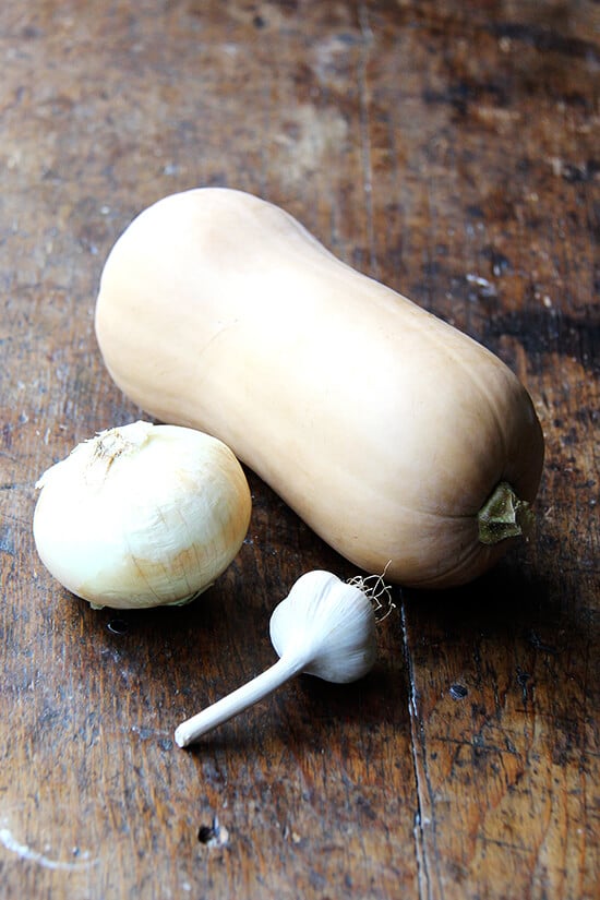 A butternut squash, onion, and garlic head on a board. 