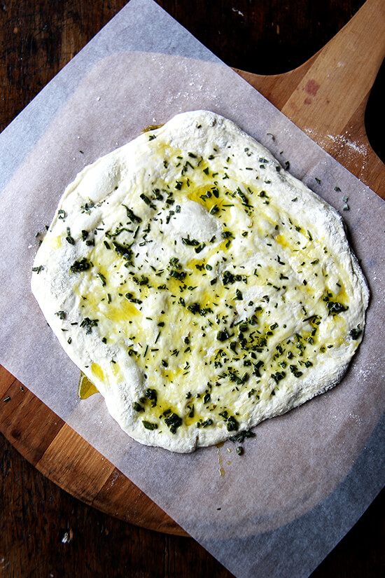 An herbed unbaked flatbread on a sheet of parchment on a baking peel. 