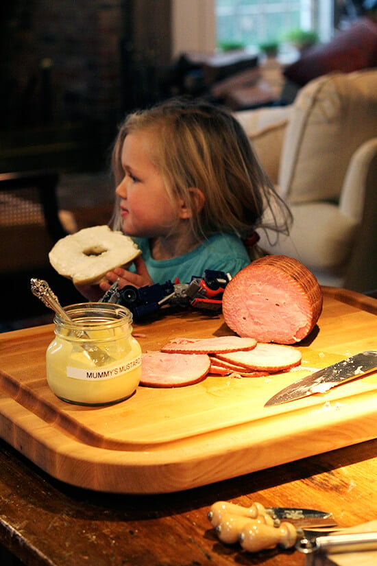 Ella eating a bagel next to a board with ham and mustard sauce. 