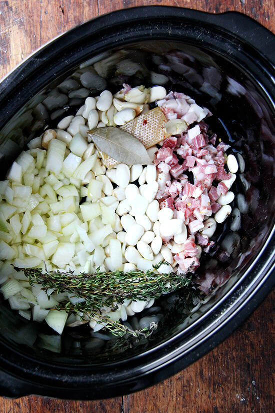 A crockpot filled with onions, beans, pancetta, bay leave, thyme, and a parmesan rind.