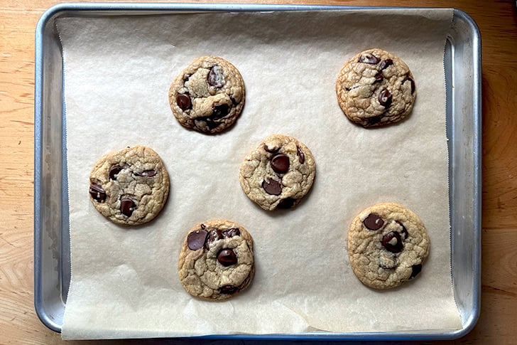 Six just-baked chocolate chip cookies on a parchment-lined sheet pan.