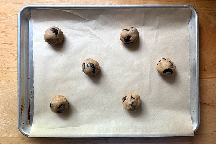 Six unbaked chocolate chip cookie dough balls on a parchment-lined sheet pan.
