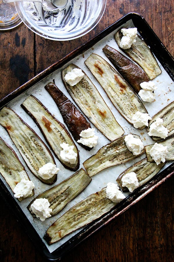 Roasted eggplant on a sheet pan with dollops of ricotta on each end, ready to be rolled.