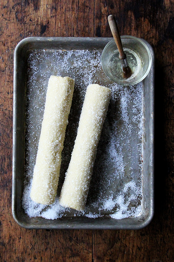 Two logs of sablé cookie dough on a small sheet pan rolled in decorative sparking sugar. 