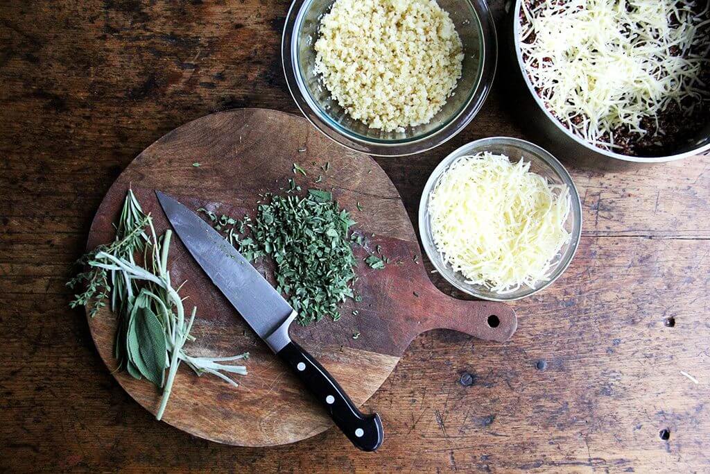 A board of chopped herbs aside bowls of bread crumbs, cheese, and quinoa. 