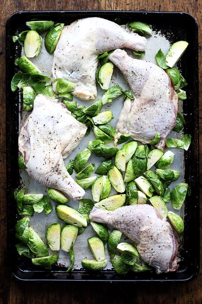 A sheet pan with uncooked chicken legs and Brussels sprouts.