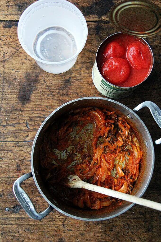 An overhead shot of a pot of cooked onions and tomato paste, a can of plum tomatoes, and a quart of water.