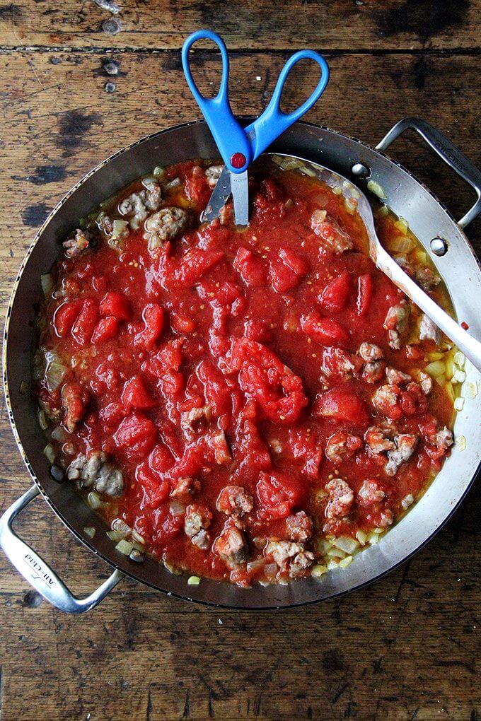 Cutting the tomatoes with scissors in a sauté pan filled with sausage, onions and tomatoes.