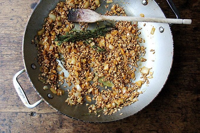 A sauté pan with onions, farro, thyme, and a bay leaf.