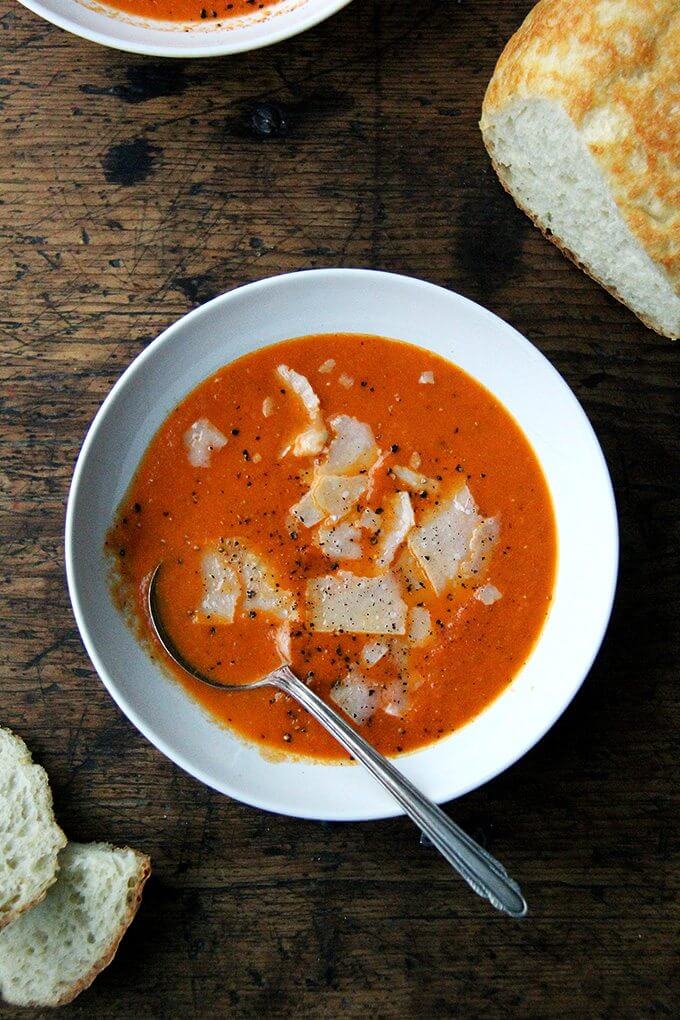 An overhead shot of a bowl of tomato soup with a spoon in it, shaved parmesan, and freshly cracked pepper.