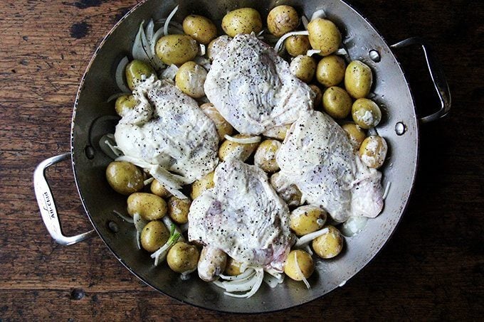 A braiser filled with potatoes, onions, chicken thighs, salt and pepper.