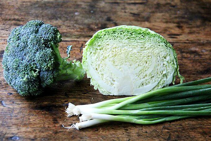 A head of broccoli, cabbage, and scallions on a board. 