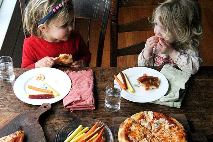 Two toddlers sitting at a table eating pizza.