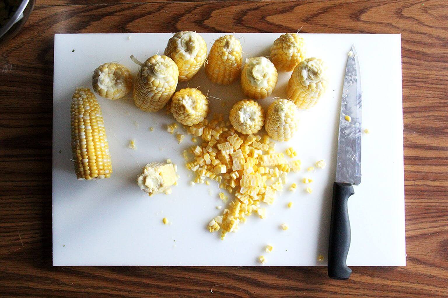 A cutting board with corn and stripped kernels and a knife.