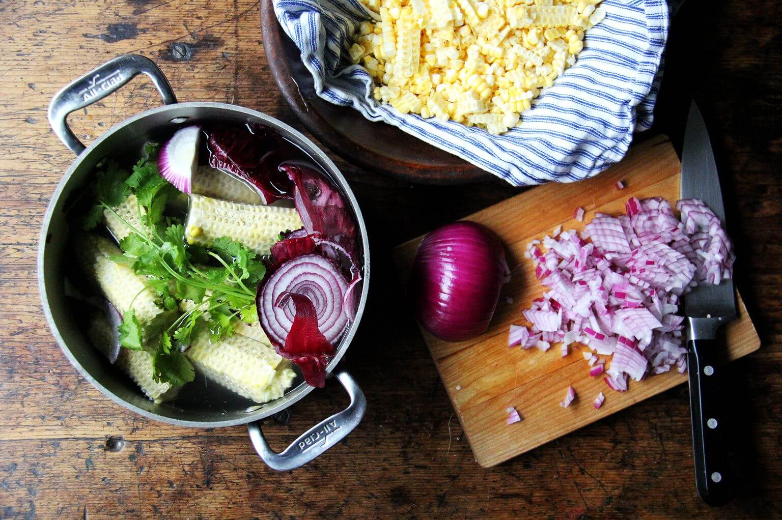 A board with a pot filled with stock ingredients, diced red onion, and stripped corn.