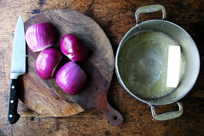 A board of halved onions aside a pot with a stick of butter in it. 