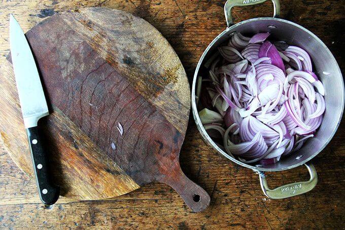 A board aside a pot with a stick of butter in it and 2 sliced onions.