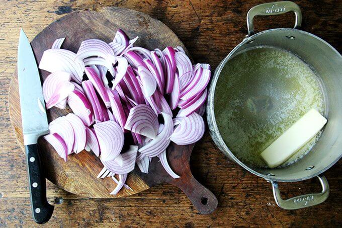 A board of sliced onions aside a pot with a stick of butter in it.