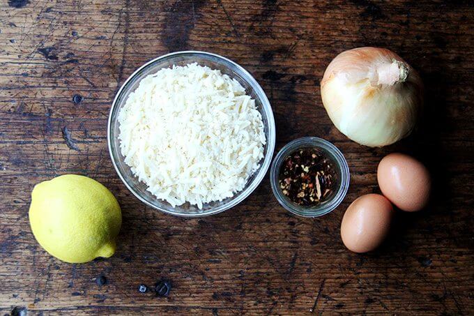 A table with a lemon, bowl of parmesan, onion, pepper flakes, and two eggs on it. 