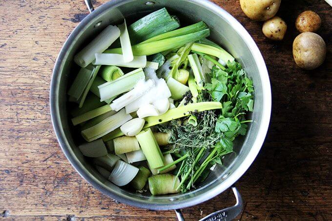 A pot of vegetable stock on a table next to a few potatoes. 