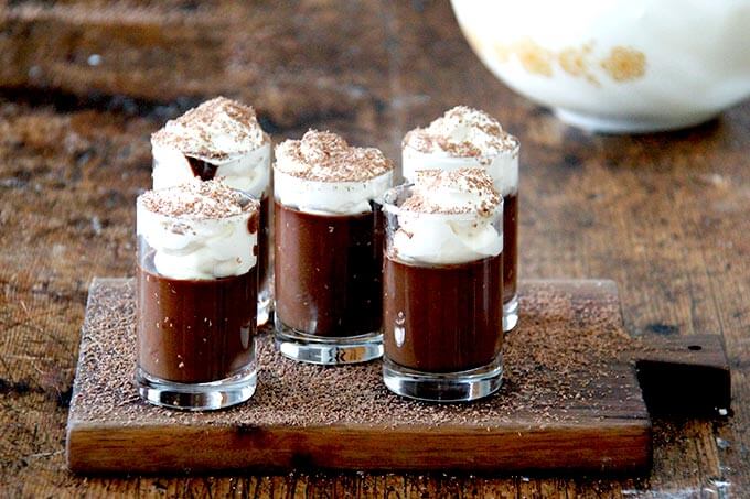 A board filled with small glasses filled with chocolate pot de creme and topped with homemade whipped cream and shaved chocolate over top.