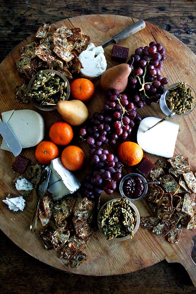 A large cheese board filled with cheese, grapes, oranges, pear, and candied pepitas.
