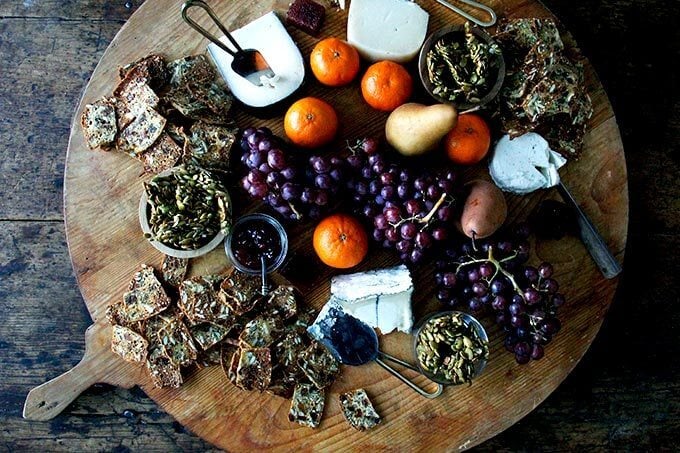 A cheese board loaded with homemade three-seed crackers, grapes, oranges, pears, and candied pepitas. 