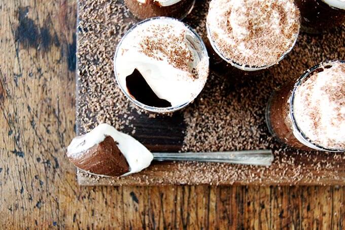 A board filled with small glasses filled with chocolate pot de creme and topped with homemade whipped cream, shaved chocolate, and a spoon.