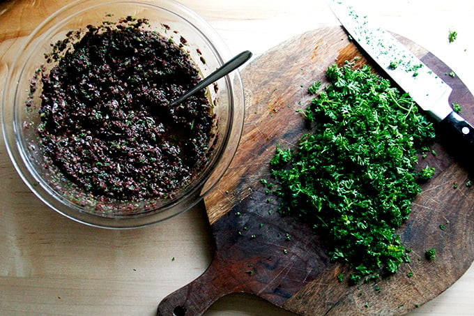 A bowl of black olive tapenade aside a chopping board with chopped parsley and a chef's knife.