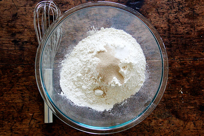 A bowl with flour, salt, sugar, and yeast aside a whisk. 