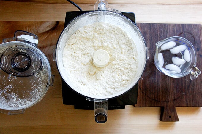 A counter with a food processor filled with dry ingredients pulsed with butter.