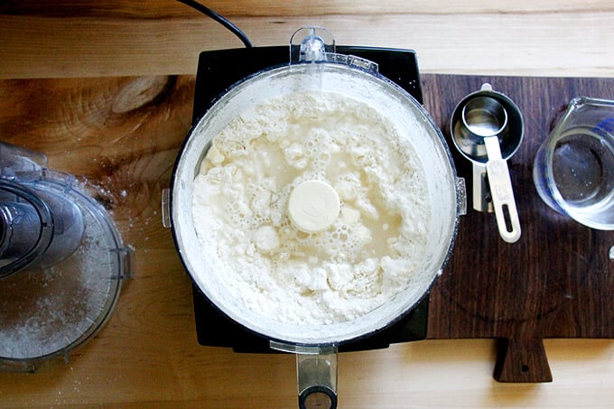 A counter with a food processor filled with dry ingredients pulsed with butter and ice water added. 