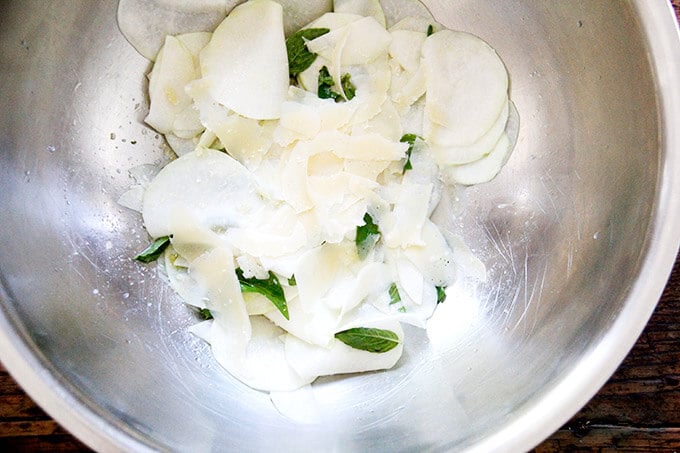 A large bowl of thinly shaved kohlrabi, salt, olive oil, vinegar, herbs, and parmesan.